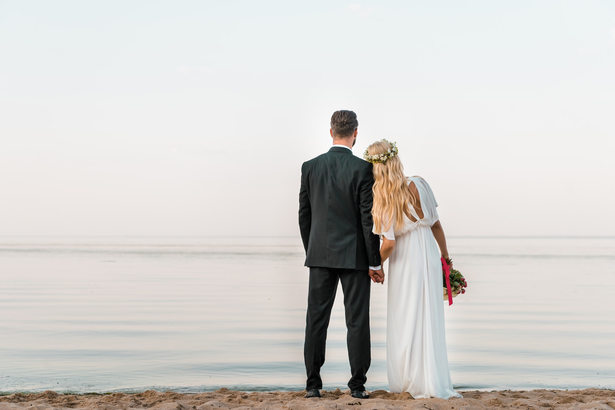 back view of wedding couple standing on beach with wedding bouquet and looking at sea